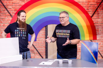 Michael Büker steht an einem Experimentiertisch neben dem Moderator Elton in einem Studio, das mit Regenbögen dekoriert ist.
